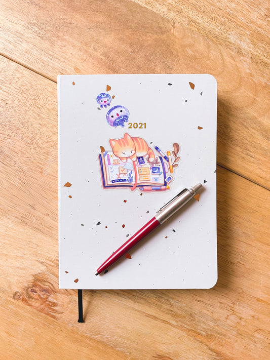Transparent vinyl kitten journal sticker: orange cat reading with “2021” and confetti dots