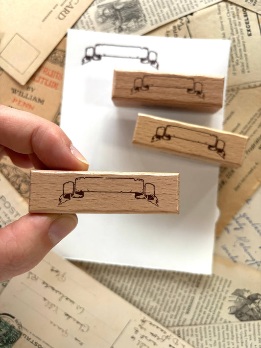 Rubber scroll banner stamp on wooden block held by hand, black outlined scroll design