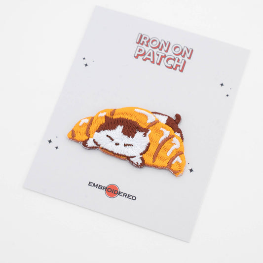 Iron-on cat food patch: curled cat sleeping on orange croissant slice, embroidered detail
