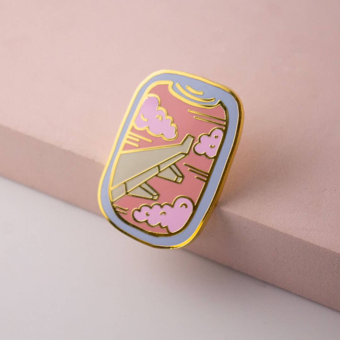 Enamel airplane window pin with pink clouds and gold outline, tilted on pink surface