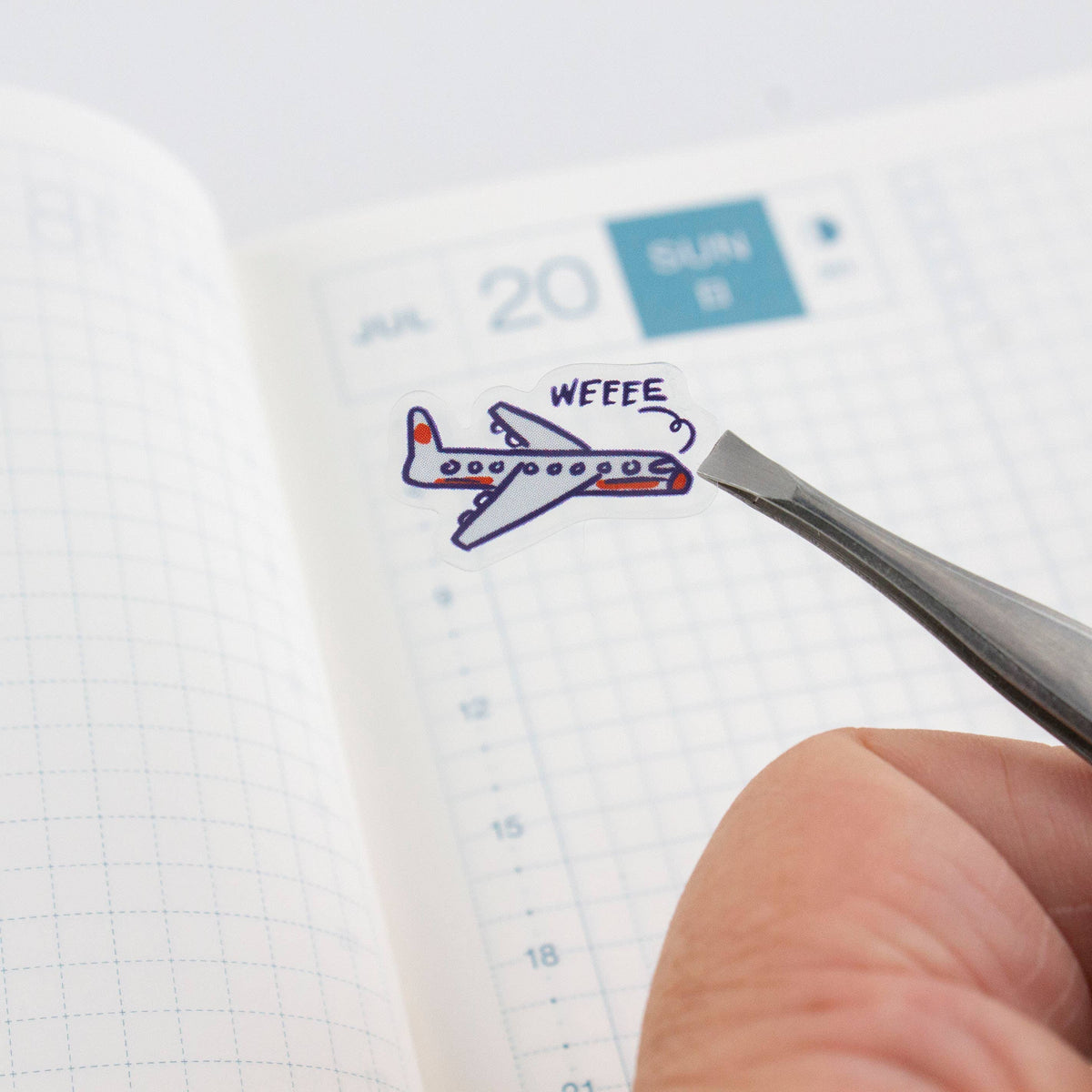 Clear planner sticker sheet with blue plane sticker being lifted by tweezers