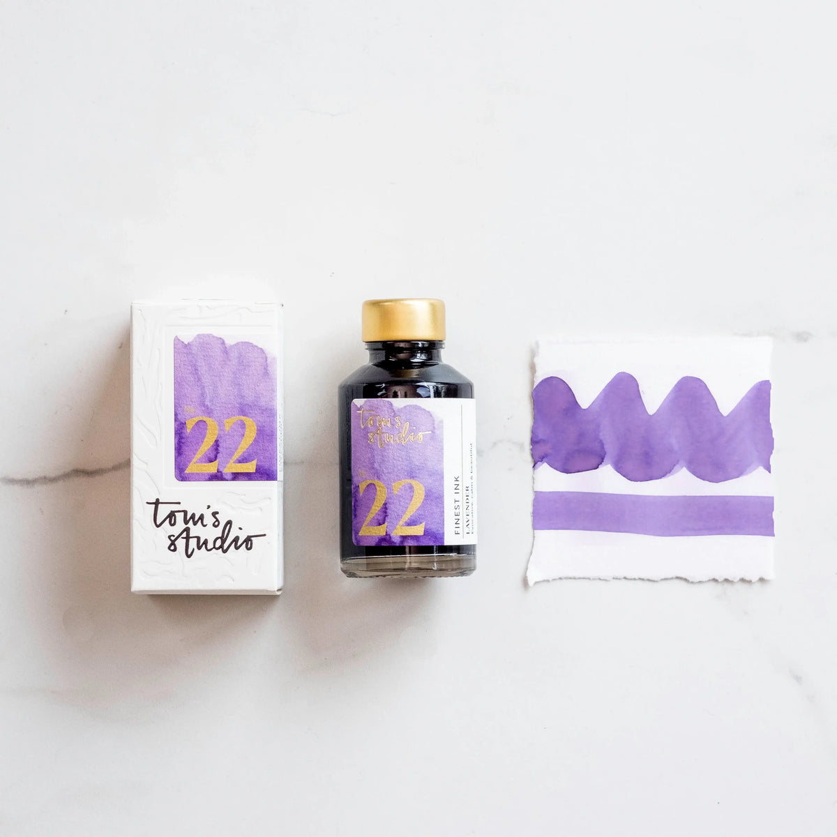 Lavender fountain pen ink bottle with gold cap and label, plus sample ink swatches