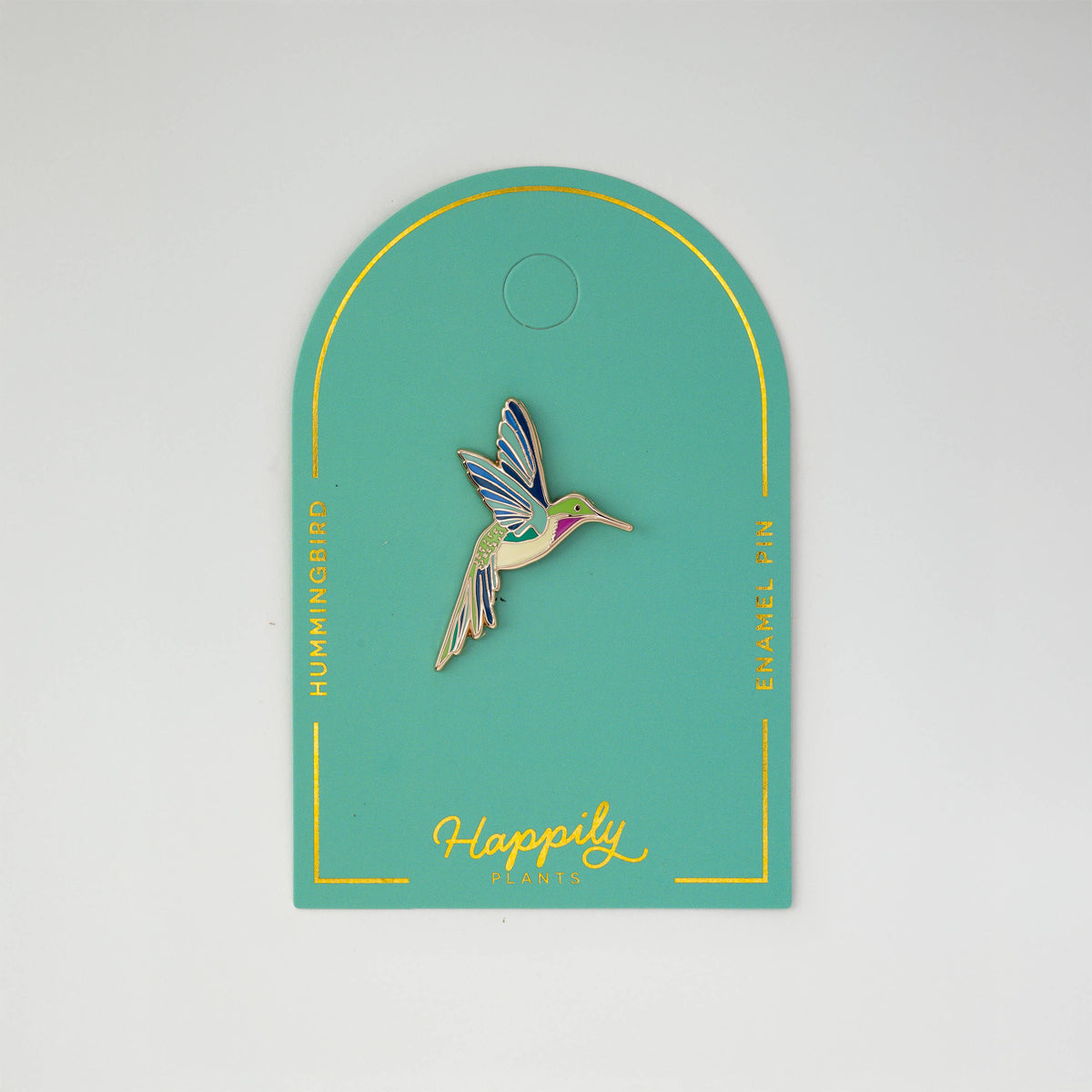 Hummingbird enamel pin centered on teal card backing with gold outline.