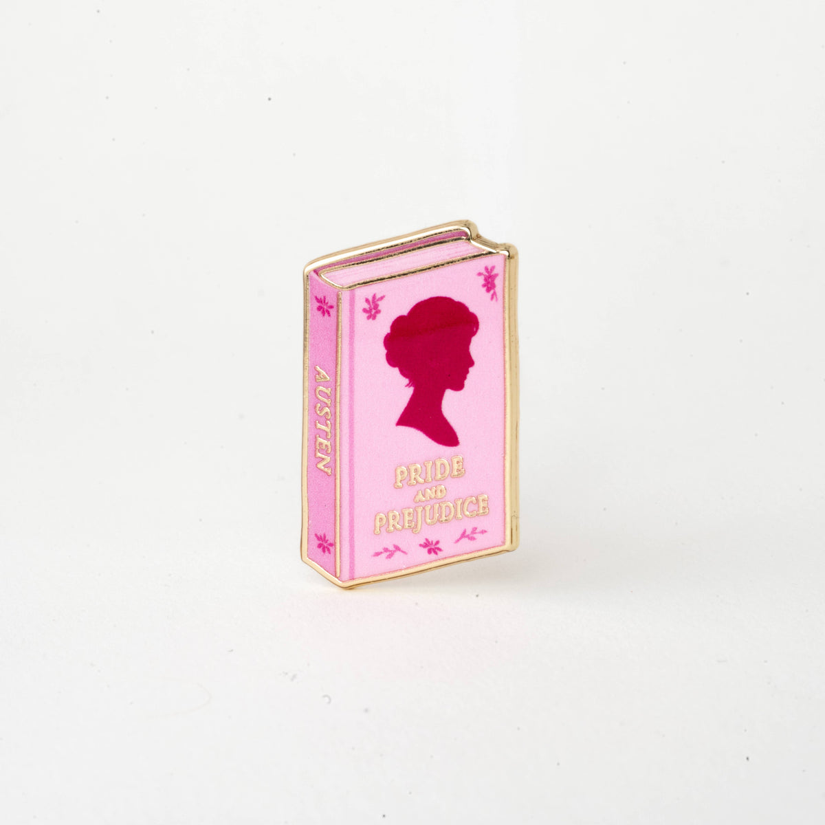 Pink book-shaped enamel pin with red silhouette and text 'Pride and Prejudice'.