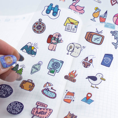Hand holding clear sticker sheet with travel icons including binoculars, flamingo, and TV