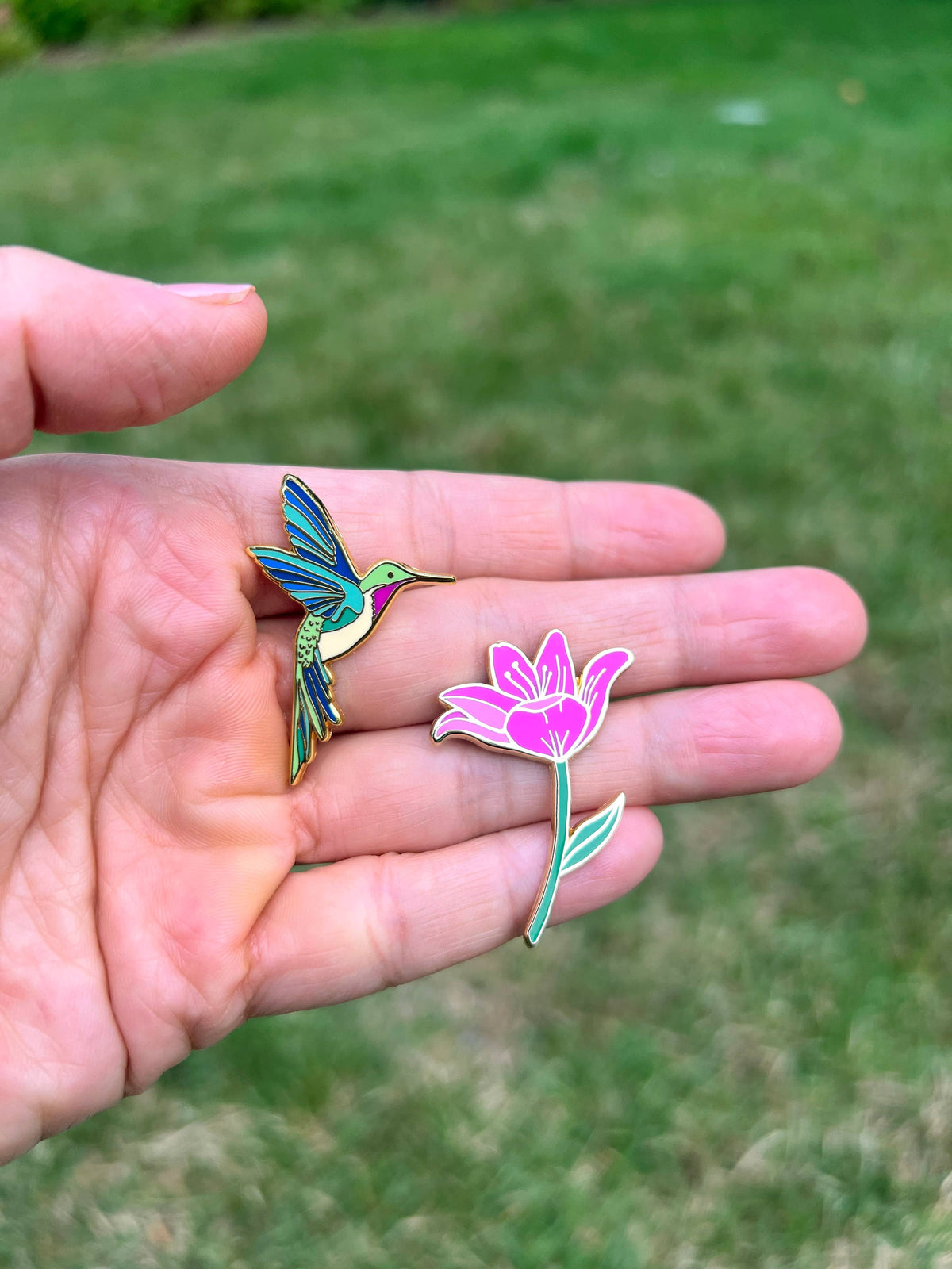 Hummingbird enamel pin held in hand with another floral pin beside it.