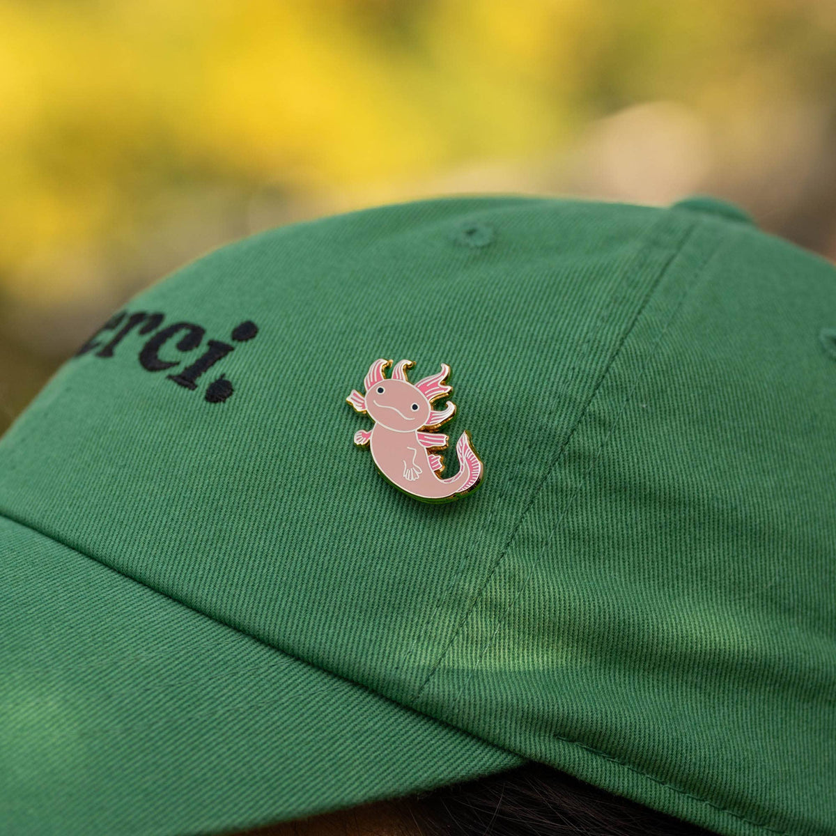 Pink axolotl enamel pin on green baseball cap, gold outline and curled tail