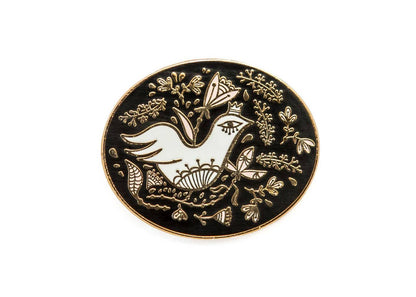 Close-up of an oval enamel pin featuring a white bird and gold floral details on black