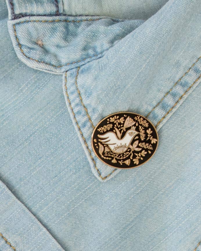 Oval floral enamel pin with a white bird and gold botanical details on a black field