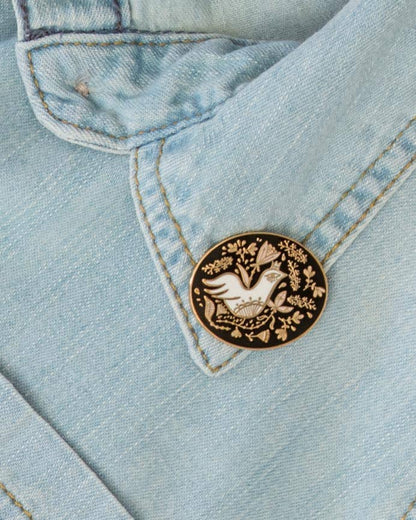Oval floral enamel pin with a white bird and gold botanical details on a black field