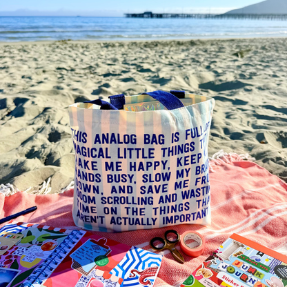 Canvas tote bag with blue handles, printed "ANALOG BAG" quote in dark lettering.