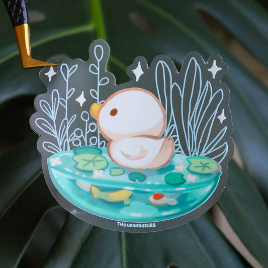 Transparent vinyl sticker with cartoon orange-and-white creature floating in teal pond