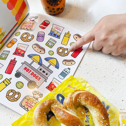 Food sticker book open to a hot dog-themed page with a hand placing stickers