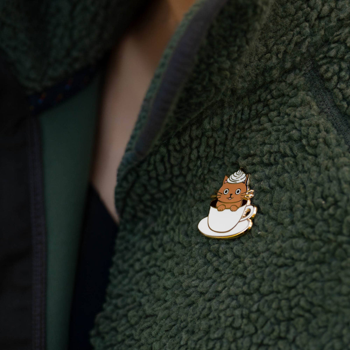 Cat latte enamel pin on dark green fleece jacket