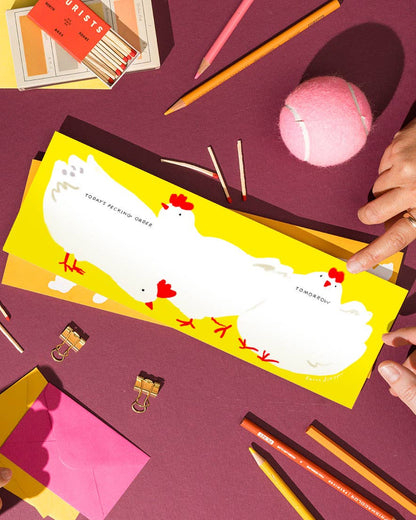 Weekly desk planner pad with two white chicken illustrations on a yellow page, shown at an angle