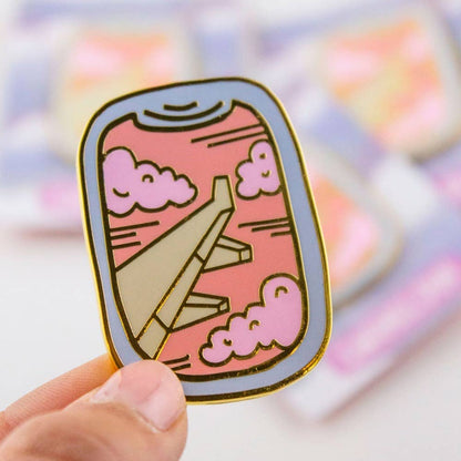 Enamel airplane window pin with pink clouds and gold outline, pin held in hand
