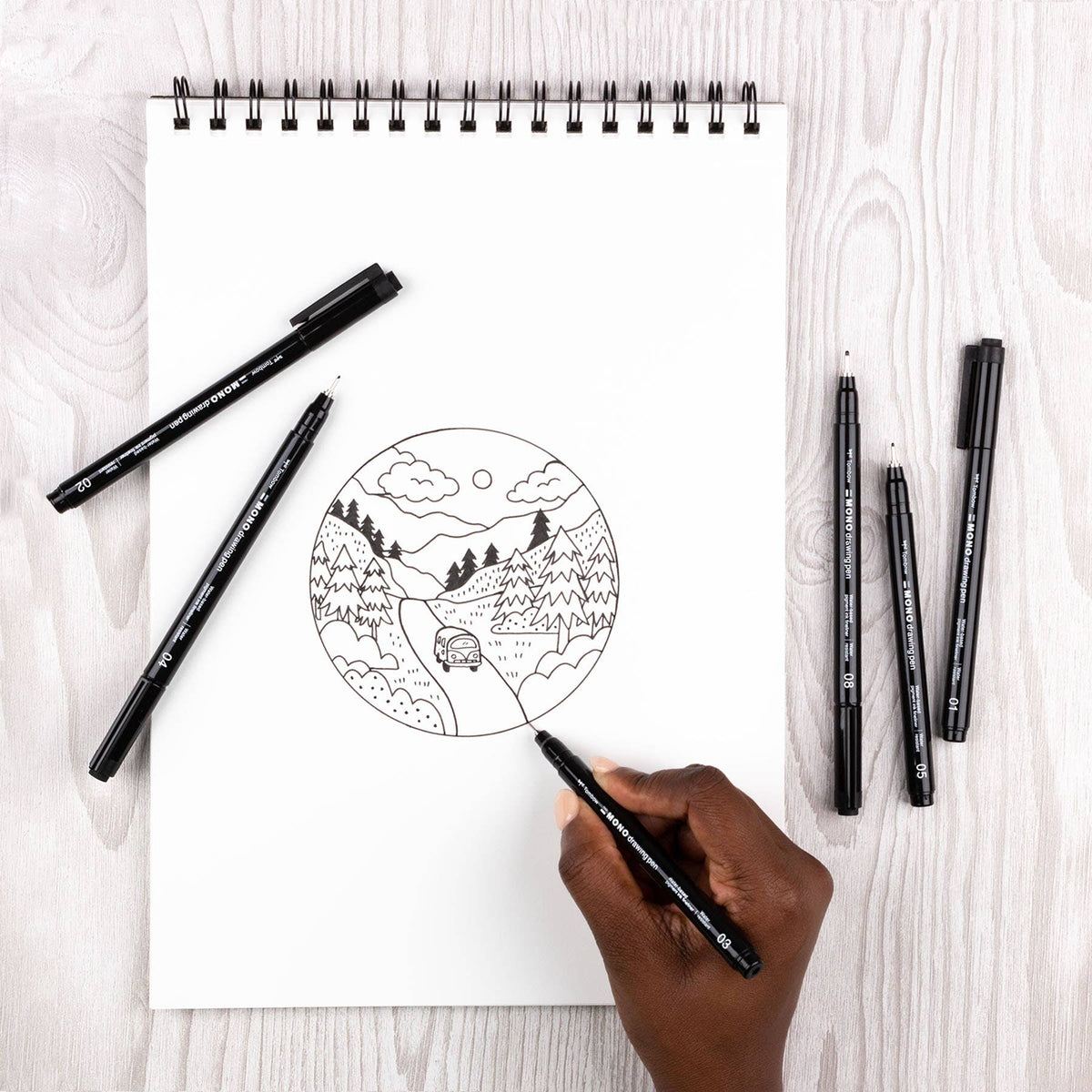 Set of black fine-tip drawing pens beside a spiral sketch on a white pad