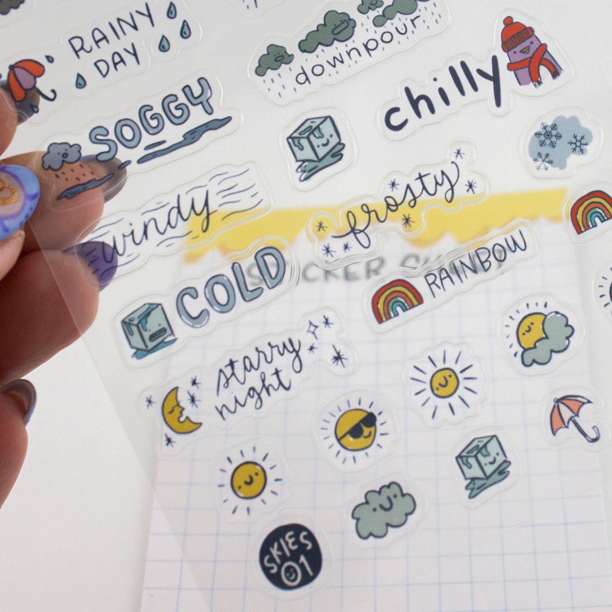 Hand holding weather sticker sheet with rainy day, soggy, windy, cold, and rainbow icons