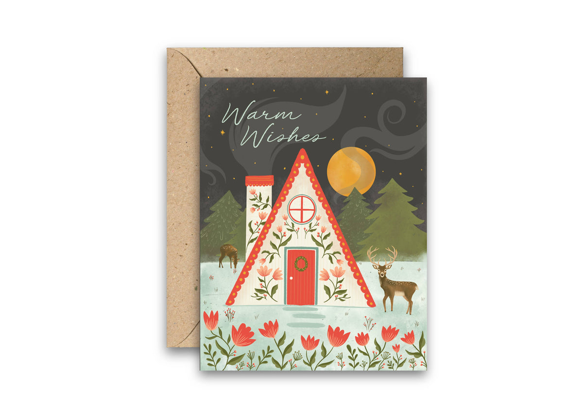 Woodland Christmas greeting card with a decorated cabin, deer, trees, and matching kraft envelope