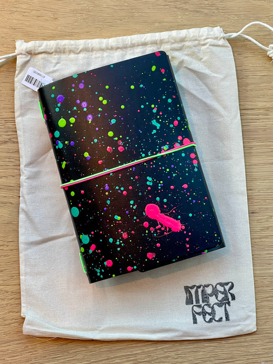 Dark speckled notebook cover with neon paint splatter and pink raven, in a white drawstring bag