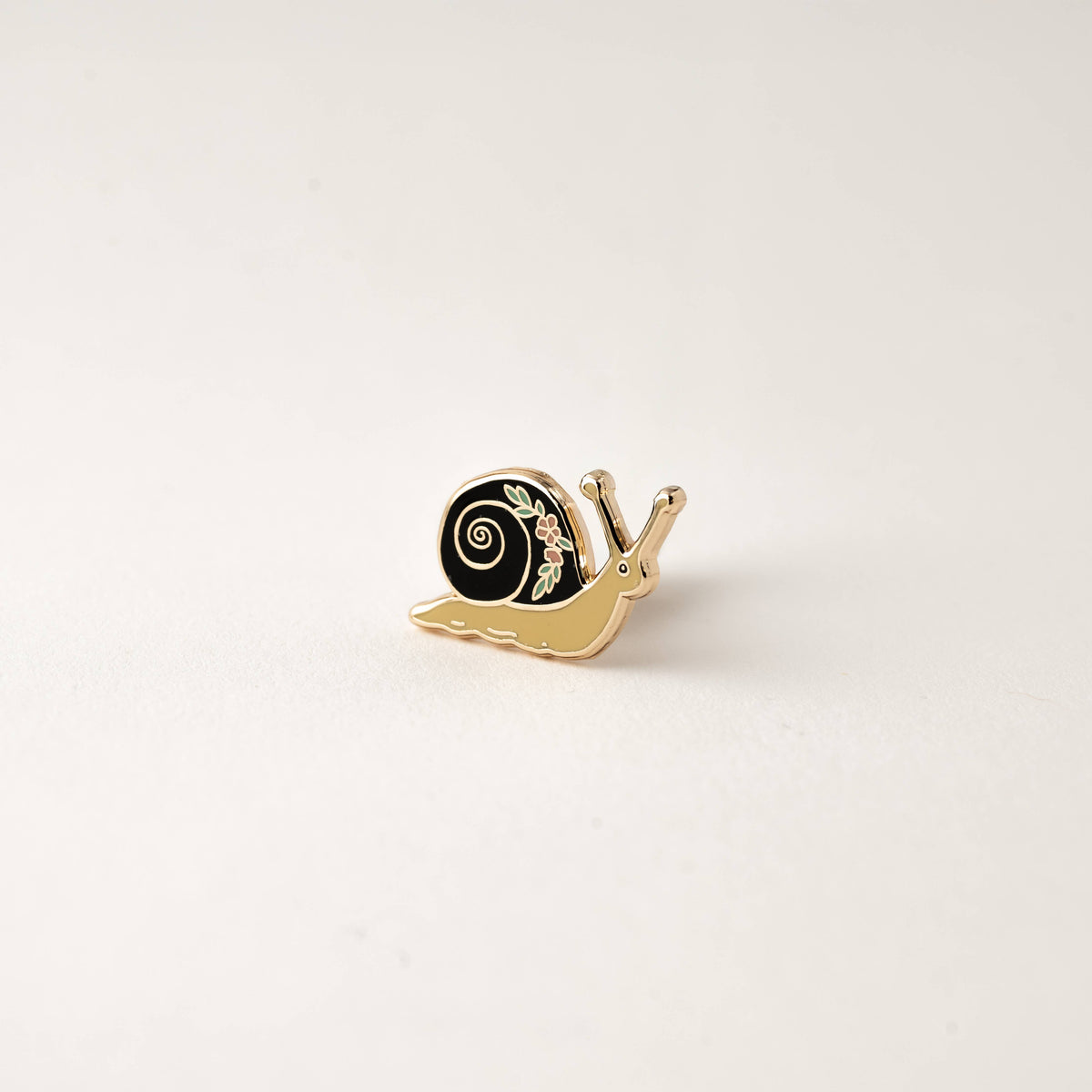 Small garden snail enamel pin with black spiral shell, gold-tone outline and flower detail