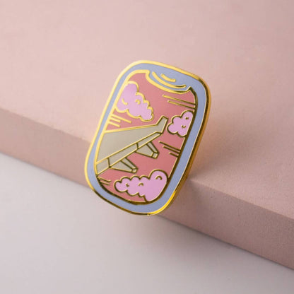 Enamel airplane window pin with pink clouds and gold outline, tilted on pink surface