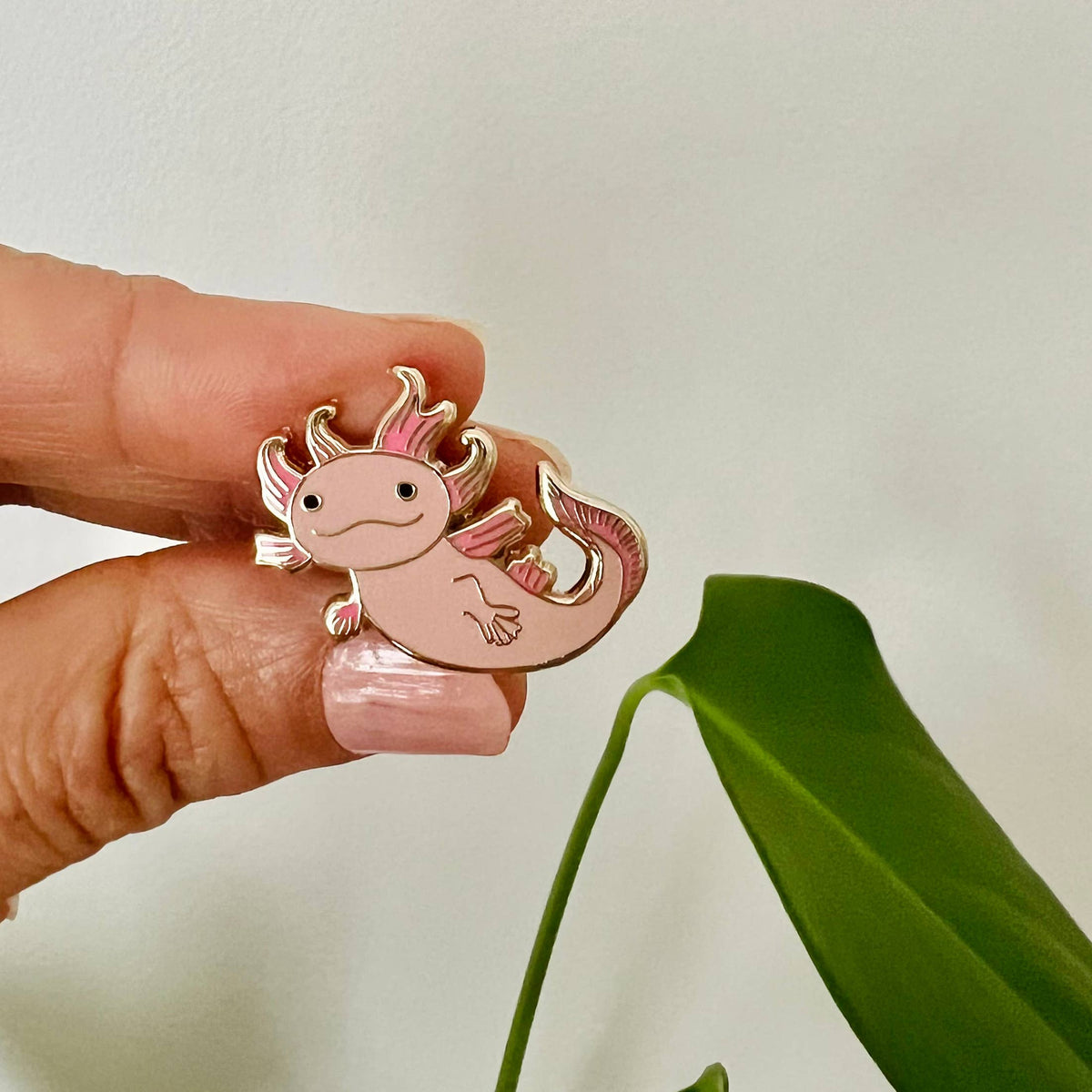 Pink axolotl enamel lapel pin held between fingers, gold outline and curled tail