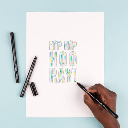 Black fine-tip drawing pen in hand adding “RAY!” lettering on a small card