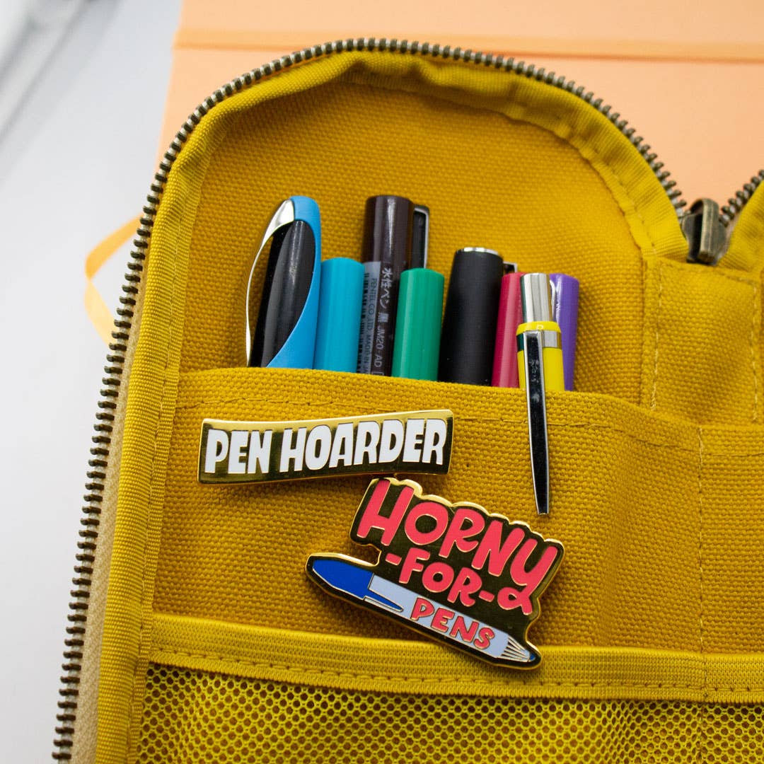 Two enamel pins on yellow pouch: 'Pen Hoarder' and 'Horny for Pens'