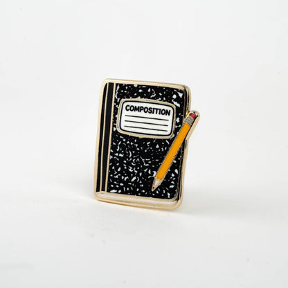 Enamel composition notebook pin with black notebook, 'COMPOSITION' label, and orange pencil