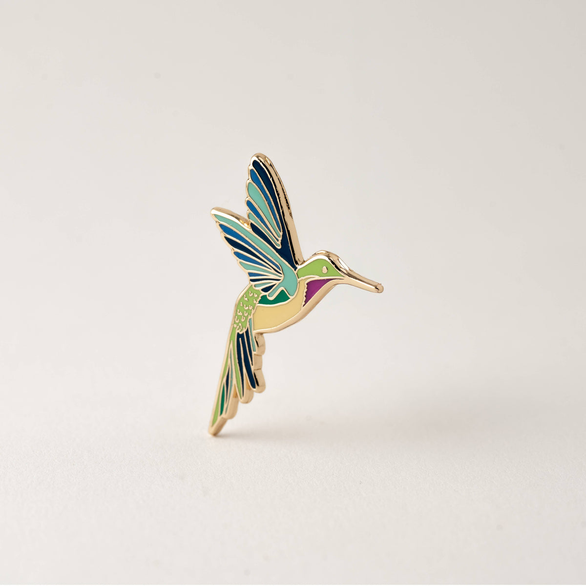 Isolated hummingbird enamel pin with blue-and-green wings and gold-tone accents.