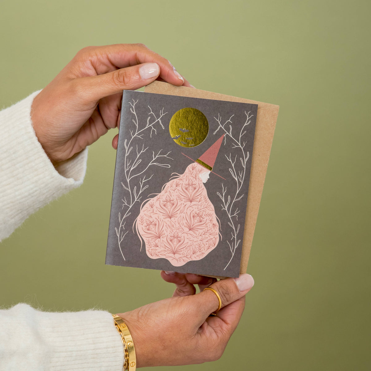 Witch greeting card with a patterned pink dress and gold moon, held in two hands