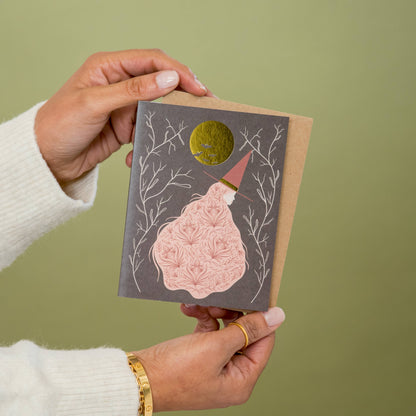 Witch greeting card with a patterned pink dress and gold moon, held in two hands