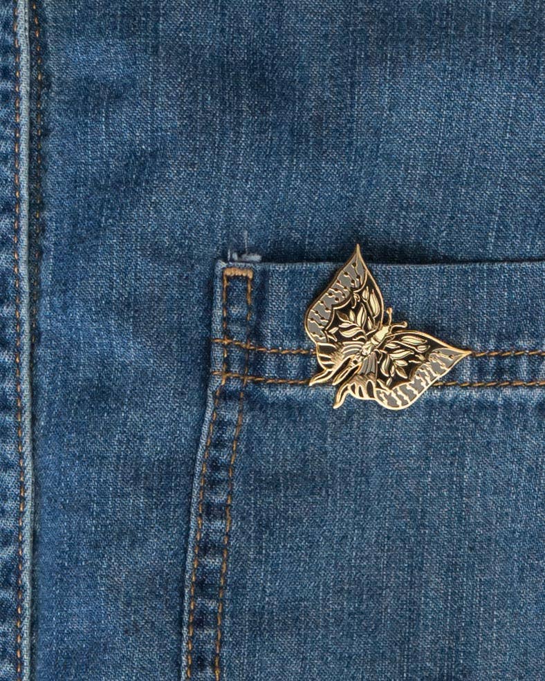 Moth enamel pin with gold outlines and pale blue and black wings, attached to a denim pocket.