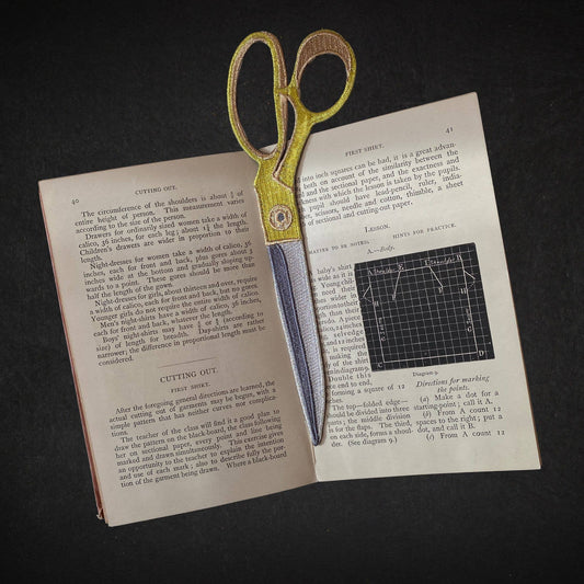 Embroidered scissors fabric bookmark with yellow-and-brown edging, lying atop open pages