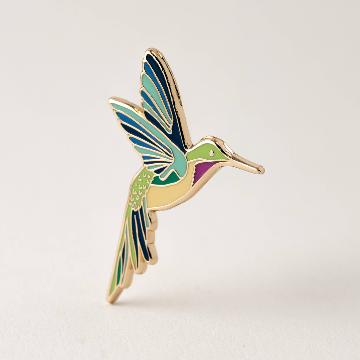 Hummingbird enamel pin with blue wings, green body, and gold-tone beak and legs.