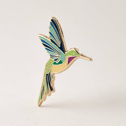 Hummingbird enamel pin with blue wings, green body, and gold-tone beak and legs.