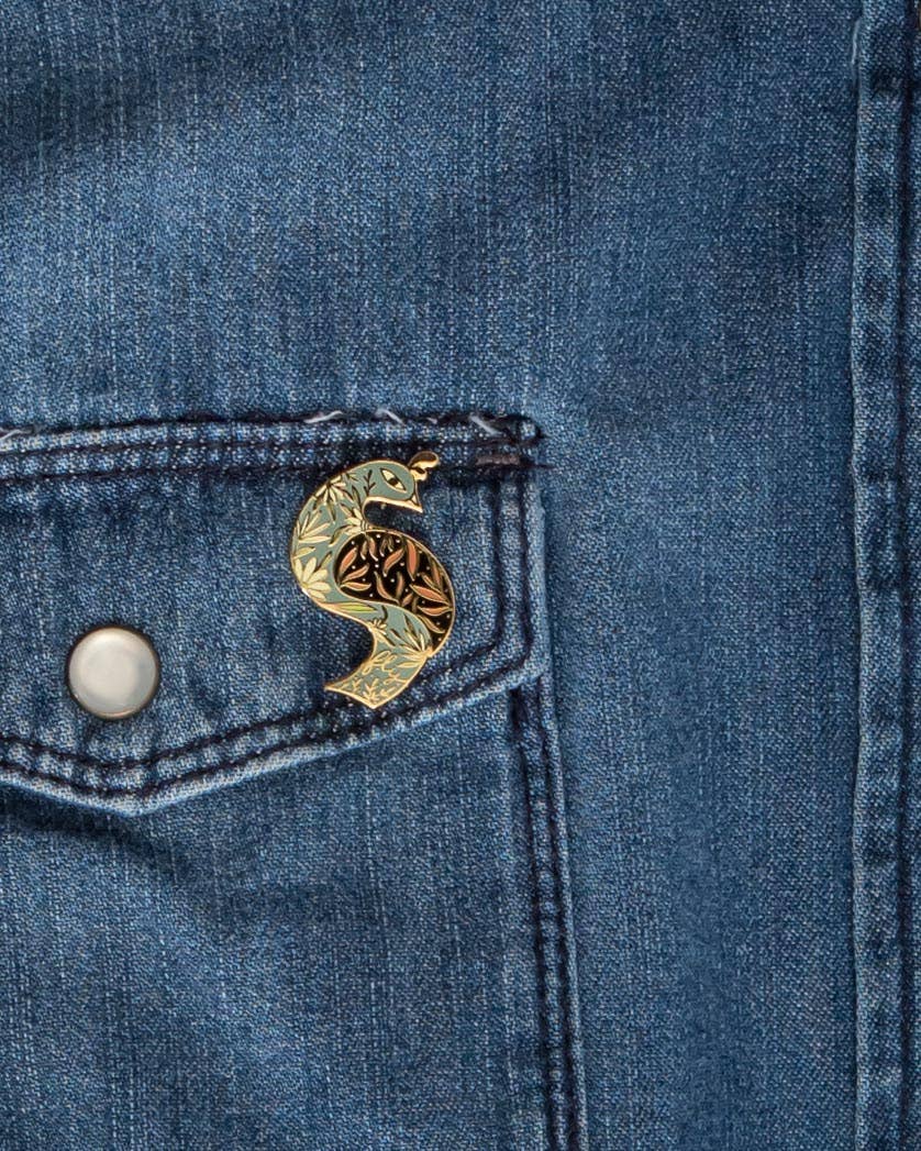 Floral bird enamel pin attached to a denim pocket, with gold-tone outlines and patterned details.