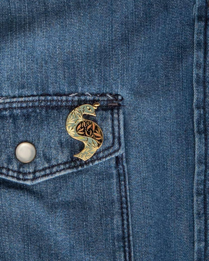 Floral bird enamel pin attached to a denim pocket, with gold-tone outlines and patterned details.