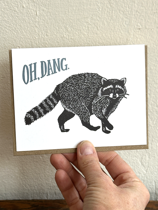 Raccoon letterpress card front held in hand, “OH, DANG.” text with black raccoon art