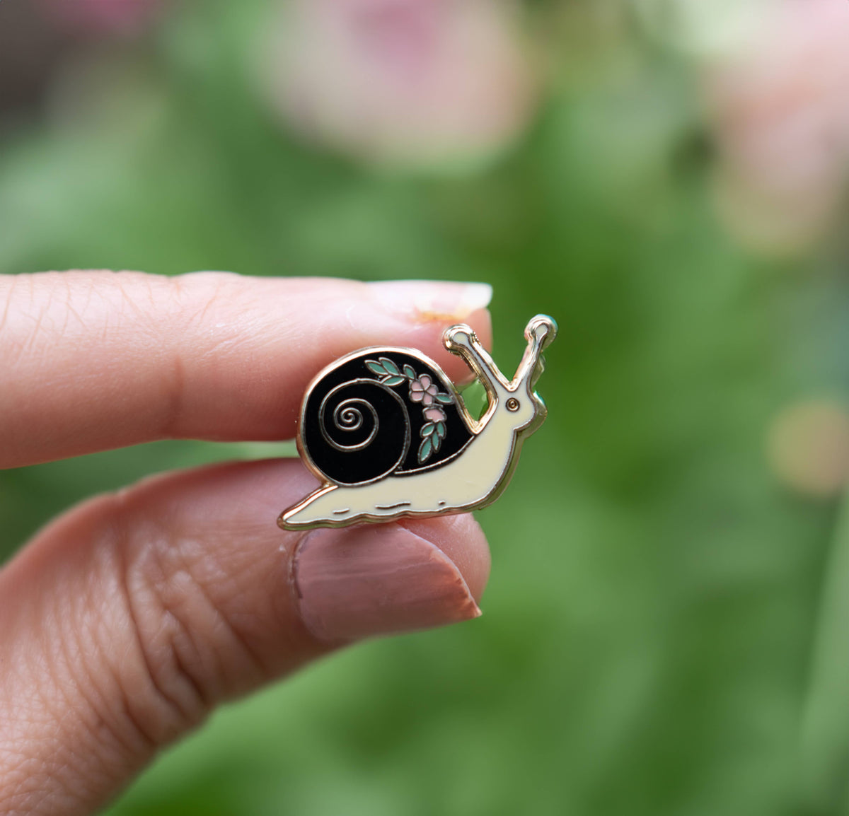 Snail enamel lapel pin in hand, black swirl shell with flower accent, gold-tone finish