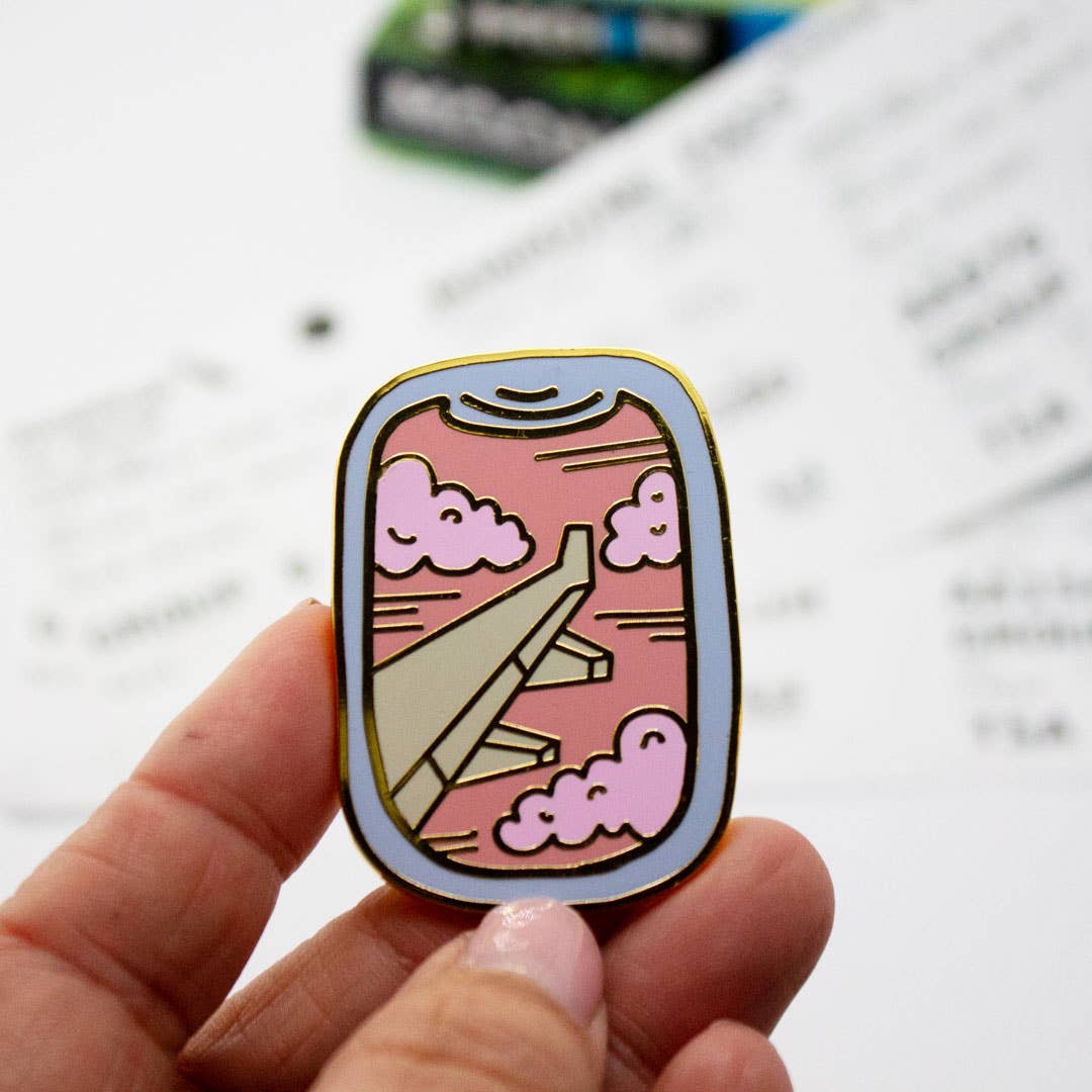 Enamel airplane window pin with pink clouds and gold outline, held in fingers