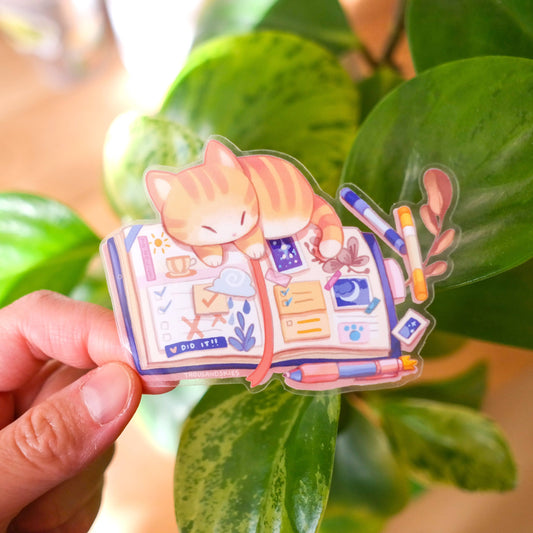 Transparent vinyl kitten journal sticker: orange cat sleeping in open notebook with pens