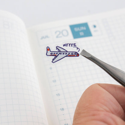 Clear planner sticker sheet with blue plane sticker being lifted by tweezers